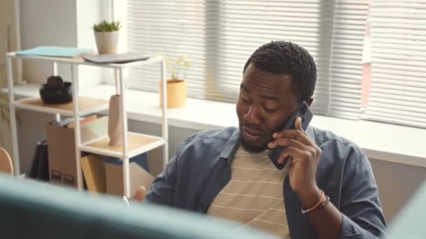 Nuori musta mies istuu työpöydällä modernissa toimistossa ja puhuu matkapuhelimella - Materiaali, video