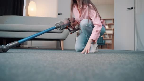Młoda Europejka w przytulnych domowych ubraniach czyści dywan z psiej sierści złotego terrier Yorkshire z bezprzewodowym odkurzaczem w mieszkaniu - Materiał filmowy, wideo