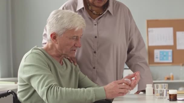 Vecchio sulla sedia a rotelle con bottiglia di pillole in mano e sua moglie che parla con il medico durante l'appuntamento in clinica - Filmati, video