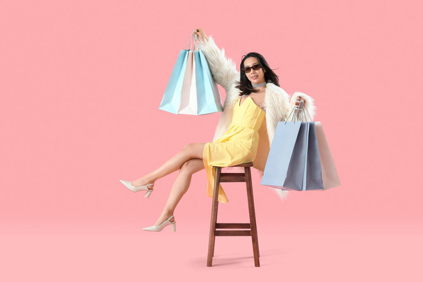 Happy beautiful young Asian woman in sunglasses with shopping bags sitting on chair against pink background - Photo, Image