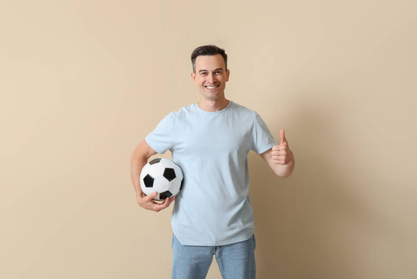 Fešák muž s fotbalovým míčem ukazující palec nahoru na béžové pozadí - Fotografie, Obrázek