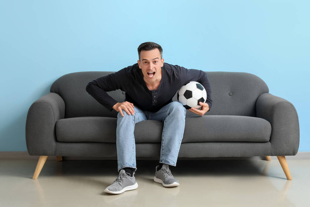 Σοκαρισμένος νεαρός άνδρας με μπάλα βλέποντας ποδόσφαιρο παιχνίδι στον καναπέ κοντά στο μπλε τοίχο - Φωτογραφία, εικόνα