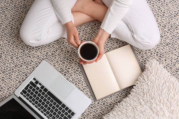Женщина с чашкой кофе, книгой и ноутбуком сидит на полу дома - Фото, изображение