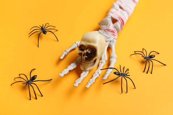 Skeletová ruka s lebkou a pavouky pro halloweenskou oslavu na oranžovém pozadí, detailní záběr - Fotografie, Obrázek