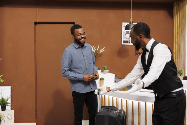 Дружелюбный афроамериканец в форме помогает гостю отеля с багажом по прибытии, посыльный улыбается и приветствует путешественника в вестибюле, помогая принести чемодан в номер - Фото, изображение