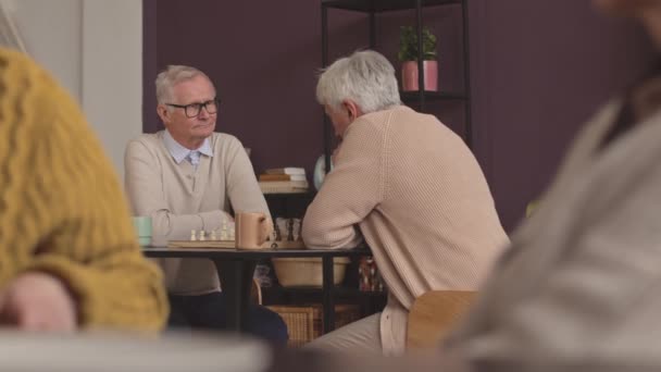 Deux hommes âgés caucasiens jouant au jeu d'échecs à table dans une maison de soins infirmiers pendant leur temps libre - Séquence, vidéo