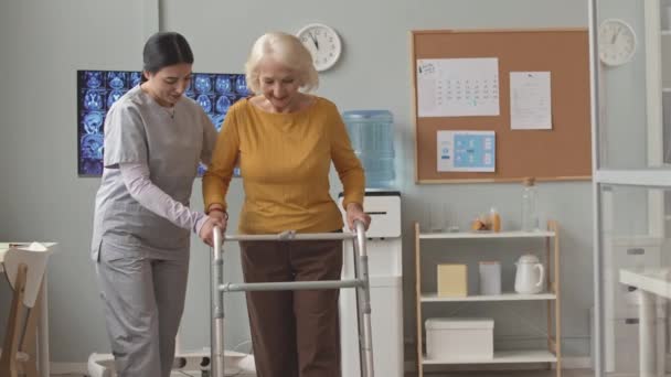 Jovem enfermeira em esfoliação ajudando a mulher idosa a caminhar ao longo do consultório de médicos apoiando-se em caminhantes - Filmagem, Vídeo