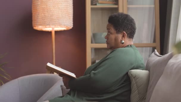 Дружелюбная молодая медсестра разговаривает с афроамериканской взрослой женщиной, читая книгу на диване в доме престарелых - Кадры, видео