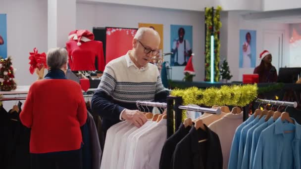 Senescent πελάτες περιήγηση μέσα από ράφια ρούχα στην εορταστική στολισμένη μπουτίκ μόδας κατά τη διάρκεια των Χριστουγέννων περίοδο των διακοπών. Ηλικιωμένο ζευγάρι ευτυχισμένη μετά την εύρεση πολύχρωμα μπλέιζερ για δώρο σε Χριστούγεννα οικογενειακή εκδήλωση - Πλάνα, βίντεο