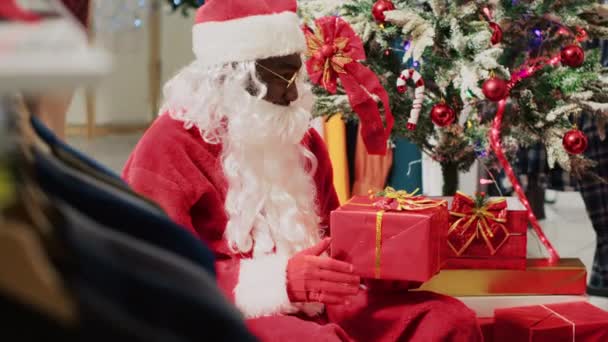 Cliente en la tienda de moda hablando con el actor vestido como Santa Claus, sentado junto al árbol de Navidad durante el evento promocional festivo. Cliente que recibe regalo del trabajador durante la temporada de vacaciones - Imágenes, Vídeo