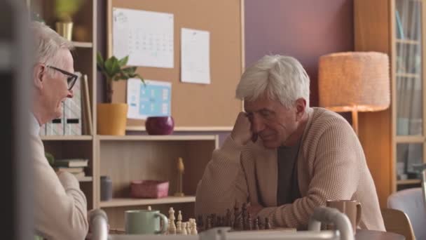 Zwei nachdenkliche kaukasische Senioren spielen Schach und unterhalten sich in der Freizeit im Pflegeheim - Filmmaterial, Video