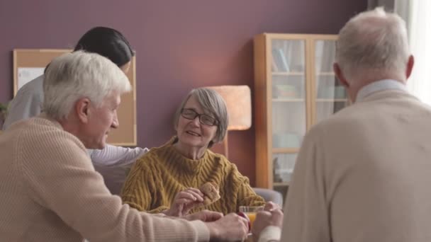 Užitečná mladá sestřička pečující o etnicky různorodé seniory sedí spolu u jídelního stolu se snídaní - Záběry, video
