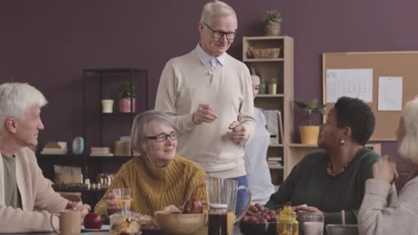 Boldog kaukázusi öregember szemüvegben beszél más idősekkel és a nővérrel az étkezőasztalnál egy otthonban. - Felvétel, videó