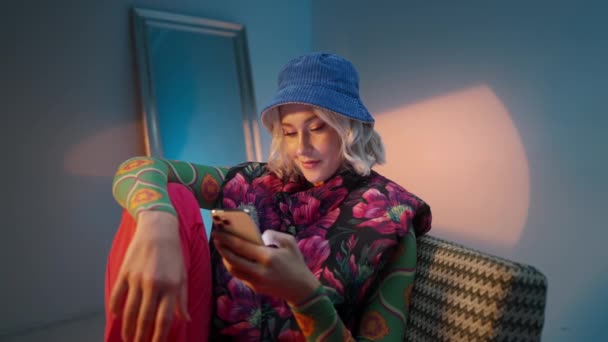 Belle jeune mannequin assise sur un fauteuil, vêtue de vêtements tendance et cherchant internet et les médias sociaux sur son smartphone dans un studio chic - Séquence, vidéo
