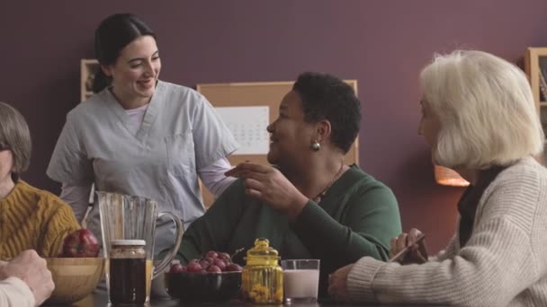 Giovane infermiera felice in scrub chatta con diversi anziani seduti in sala da pranzo in casa di cura - Filmati, video