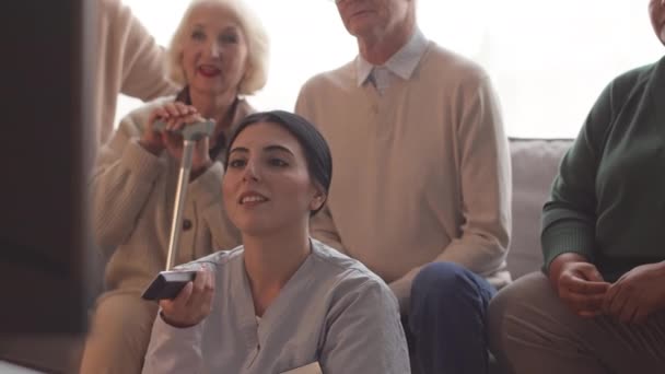 Группа веселых пожилых людей, сидящих на диване в доме престарелых и смотрящих телевизор, в то время как их молодая медсестра переключает каналы - Кадры, видео