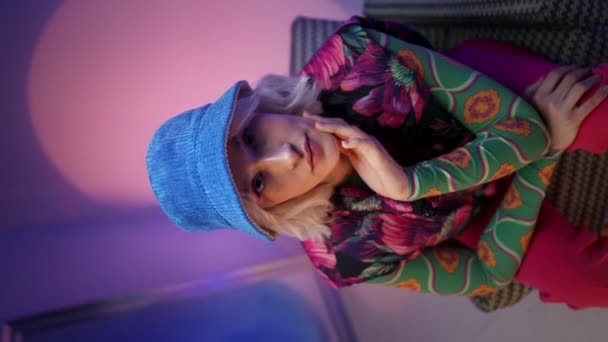Vertikální pohled na mladou stylovou modelku v klobouku, sedící v křesle a pózující před kamerou v purpurové barvě efektní studio. - Záběry, video