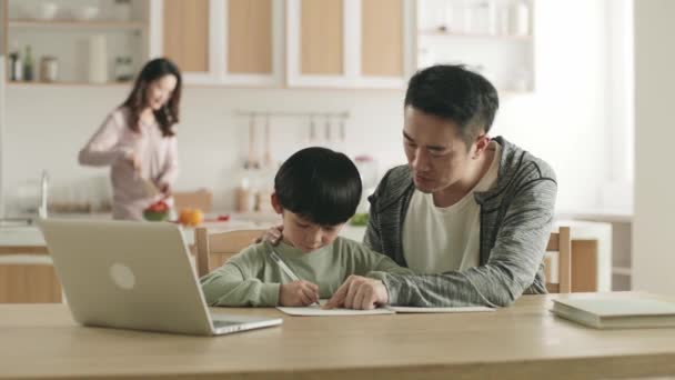 joven asiático padre ayudar hijo con homework en casa mientras madre preparación comida en cocina - Metraje, vídeo