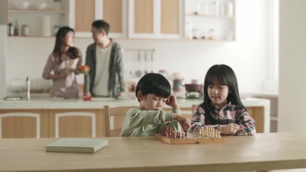twee jonge aziatische kinderen spelen thuis schaken terwijl ouders bereiden maaltijd in familie keuken - Video