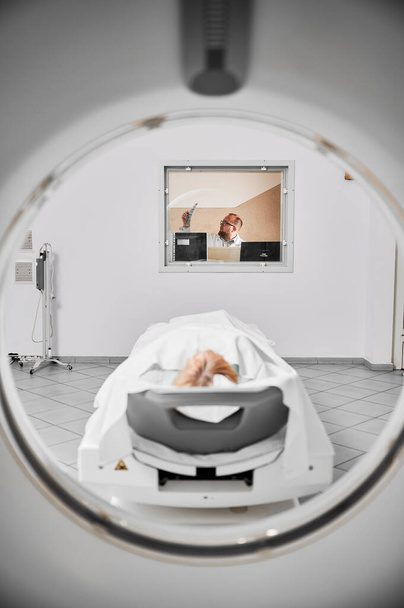 Ιατρική υπολογιστική τομογραφία ή μαγνητική τομογραφία. Γιατρός που εξετάζει τα αποτελέσματα της μαγνητικής τομογραφίας, γυναίκα ασθενής ξαπλωμένη σε καναπέ. Γιατρός με γυαλιά, κρατώντας αποτελέσματα μαγνητικής τομογραφίας, κοιτάζοντας, μελετώντας, σκεπτόμενος. - Φωτογραφία, εικόνα