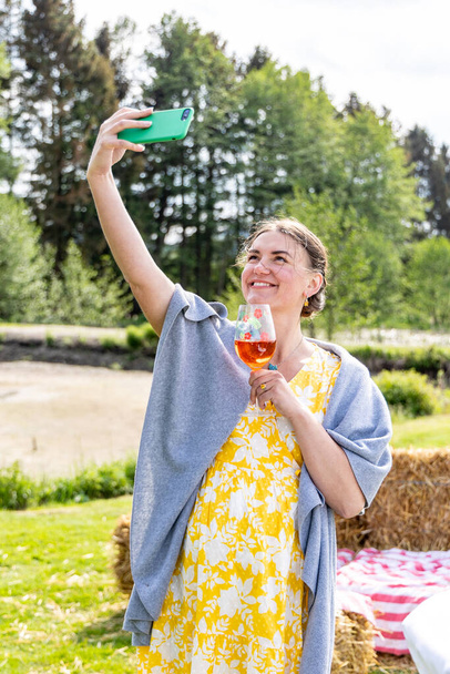 Женщина с бокалом вина в руке делает селфи на открытом воздухе - Фото, изображение