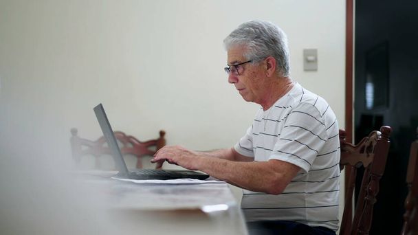 Homme âgé franc tapant sur le clavier de l'ordinateur portable à la maison. Scène domestique authentique d'une personne âgée engagée dans la technologie moderne, regardant l'écran d'ordinateur - Photo, image