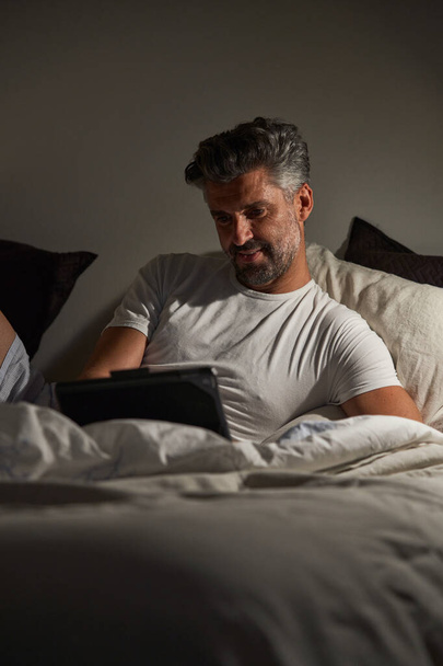 Bearded male freelancer with gray curly hair using tablet and smiling while lying on bed during work on project at night - Photo, Image