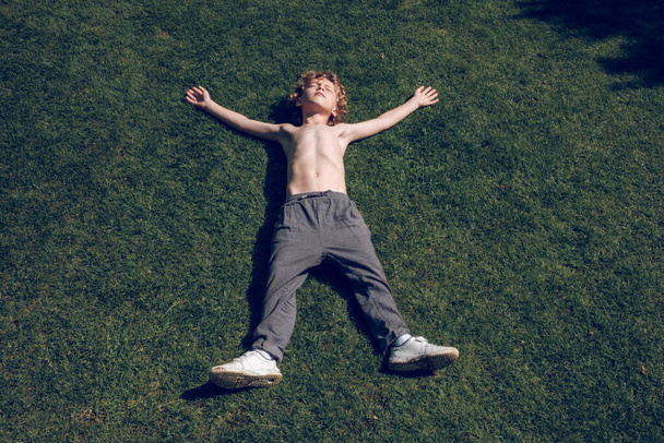 From above of carefree little kid with naked torso and curly blond hair lying on green lawn with raised arms and closed eyes in summer sunny day - Photo, Image