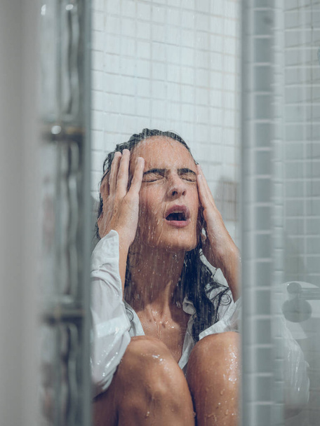 Zaklatott nő, sötét hajú, fehér, vizes ingben sír, miközben a zuhany alatt ül, csukott szemmel, és megérinti a fejét. - Fotó, kép