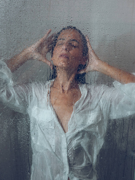 Через склянку привабливої жінки в блузці з мокрим чорним волоссям у легкій душовій кабіні з прозорими дверима у ванній - Фото, зображення