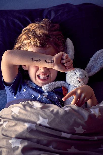 little boy covering eyes with hand. painted eyes on hand, sleepy child with bunny toy - Photo, Image