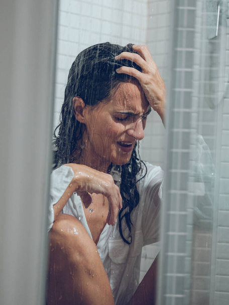 Vestida dama de pelo largo llorando en cabina de ducha mientras se sienta bajo rociar agua y tocar la frente - Foto, imagen