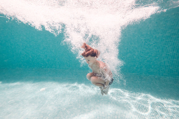 Side full body view of barefoot kid plunging in clean transparent blue pool water while jumping and embracing knees - Photo, Image