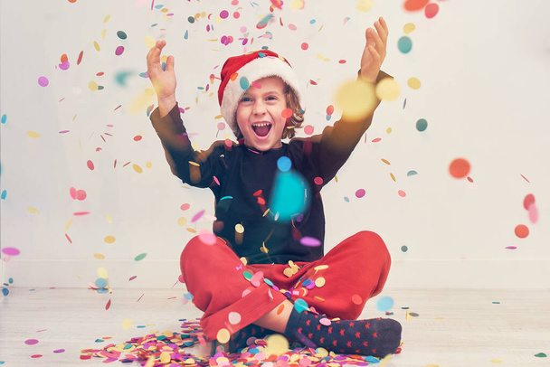 Full body of cheerful teen boy in Santa hat looking away under colorful falling confetti on white background during Christmas celebration - Photo, Image