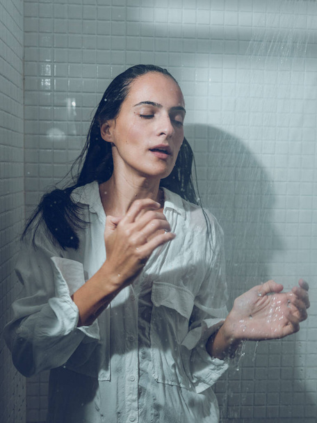 Attractive Hispanic female in white wet shirt standing with closed eyes under splashing shower in cabin in bathroom - Photo, Image