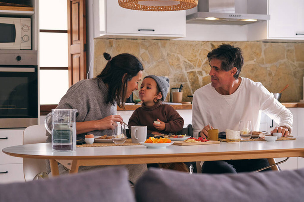 Zawartość rodziny z uroczą dziewczynką interakcji przy stole ze świeżymi plasterkami owoców patrząc na siebie w kuchni domu - Zdjęcie, obraz