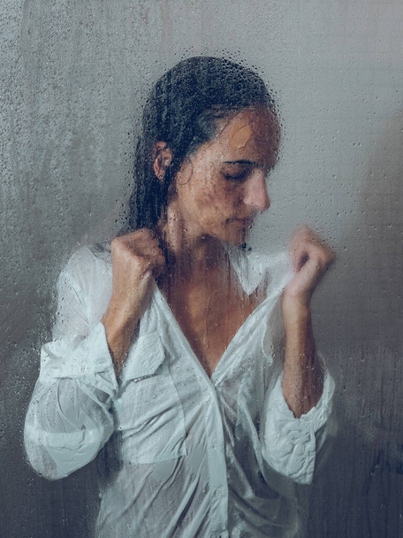Через склянку нещасної жінки в мокрому білому одязі, що стоїть в душовій кабіні з мокрими прозорими дверима під час повсякденної рутини - Фото, зображення