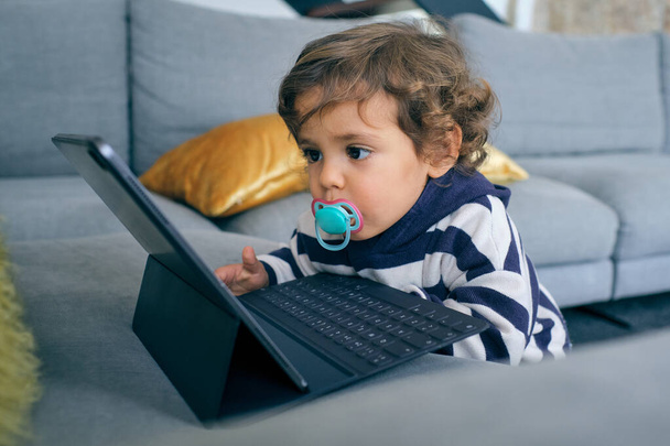 Liebenswerter kleiner Junge in lässiger Kleidung mit Schnuller, der auf dem Tablet interessante Zeichentrickfilme anschaut, während er sich zu Hause ausruht - Foto, Bild