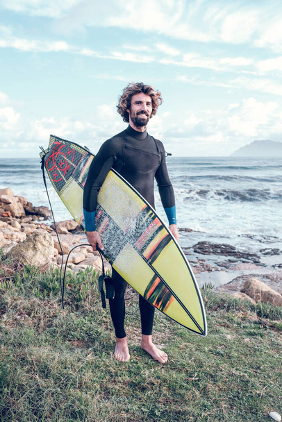 Pleine longueur de heureux jeune surfeur ethnique masculin barbu avec les cheveux bouclés en combinaison souriant tout en tenant planche de surf sur la côte rocheuse près de l'océan ondulé - Photo, image