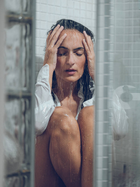 Morena solitária com olhos fechados em camisa branca molhada sentada no chuveiro de azulejos sob a água e tocando a cabeça suavemente - Foto, Imagem