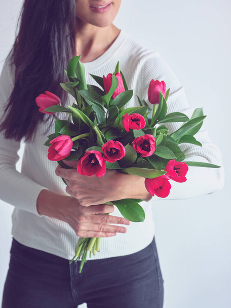 Crop señora anónima tocando y olfateando montón de tulipanes brillantes con sonrisa agradable en la cara, mientras que de pie cerca de fondo blanco - Foto, imagen