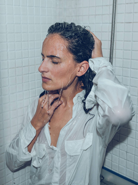 Islak gömlekli düşünceli bir kadın. Gözleri kapalı, duş kabininde su sıçratıyordu. Kafasına dokunuyordu. - Fotoğraf, Görsel