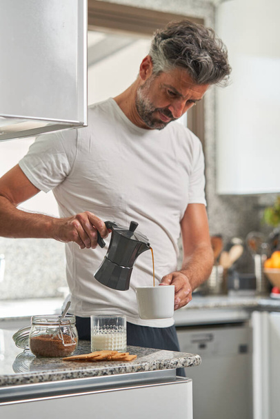 Sakallı, günlük kıyafetlerle kıvırcık saçlı erkek sabahları açık mutfakta dururken gayzer kahve makinesinden fincana lezzetli kahve döküyor. - Fotoğraf, Görsel