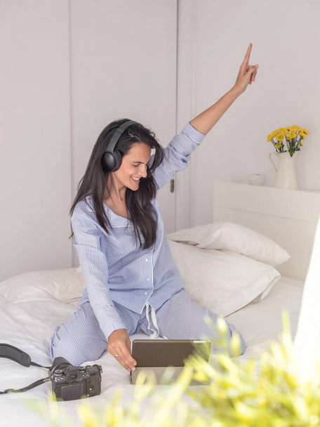 Zachwycająca kobieta w słuchawkach siedząca na łóżku i gestykulująca podczas słuchania muzyki z tabletu podczas porannej przerwy - Zdjęcie, obraz