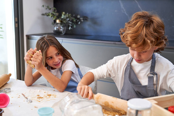 Entzückend aufmerksamer kleiner Junge mit lockigem blondem Haar in Schürze Teig kneten mit Nudelholz sitzt am Tisch neben lächelnder Schwester bei der Teigzubereitung - Foto, Bild