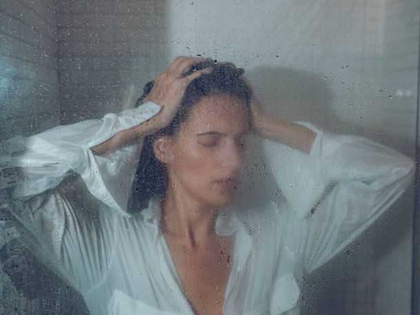 Sad sopping lady with closed eyes in white shirt touching hair behind transparent shower curtain - Photo, Image