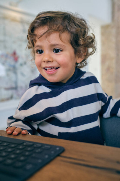 Симпатичный маленький мальчик в полосатой рубашке сидит за деревянным столом и смотрит мультфильм на нетбуке, отдыхая дома - Фото, изображение