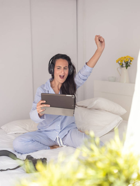 Cheerful brunette in sleepwear sitting on bed and wearing headphones while raising arm and looking at tablet in hand - Photo, Image