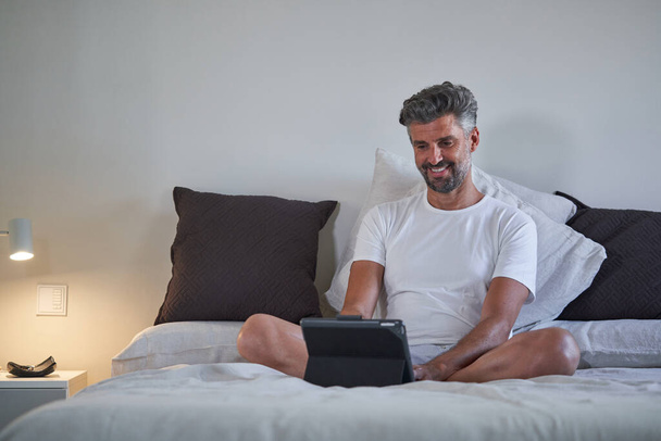 Volwassen mannelijke ondernemer in slaapkledij glimlachen en kijken naar het scherm van de tablet tijdens het werken aan project op afstand in de slaapkamer thuis - Foto, afbeelding
