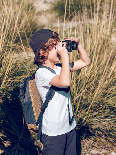 Niño rubio en ropa casual con mochila observando el campo a través de binoculares mientras está de pie cerca de la ladera de una colina cubierta de hierba en el soleado día de fin de semana en Mallorca, España  - Foto, Imagen